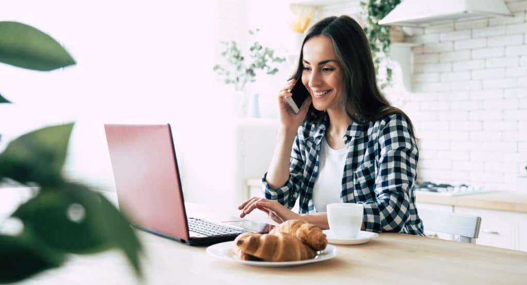 Beautiful young modern casual woman is using laptop and phone on kitchen at home. Working online.